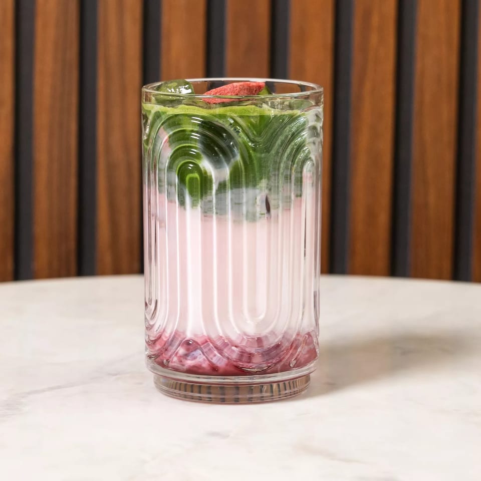 Layered pink and green drink in a textured glass with a red garnish on a marble table.