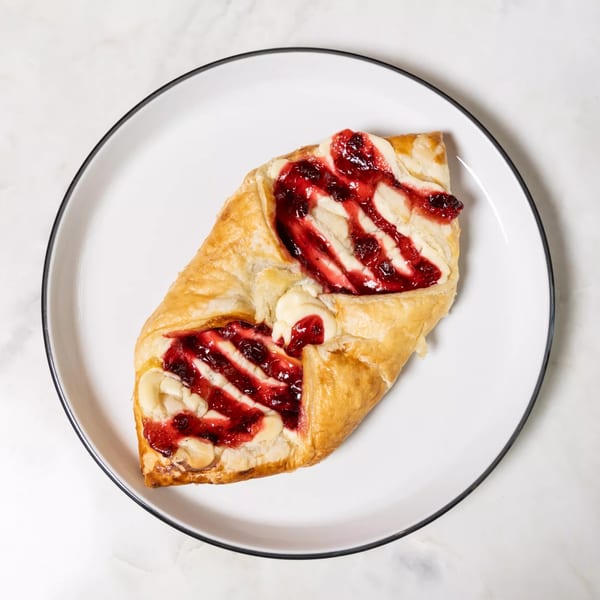 A golden-brown pastry with cream cheese and red berry jam drizzled on top, on a white plate.