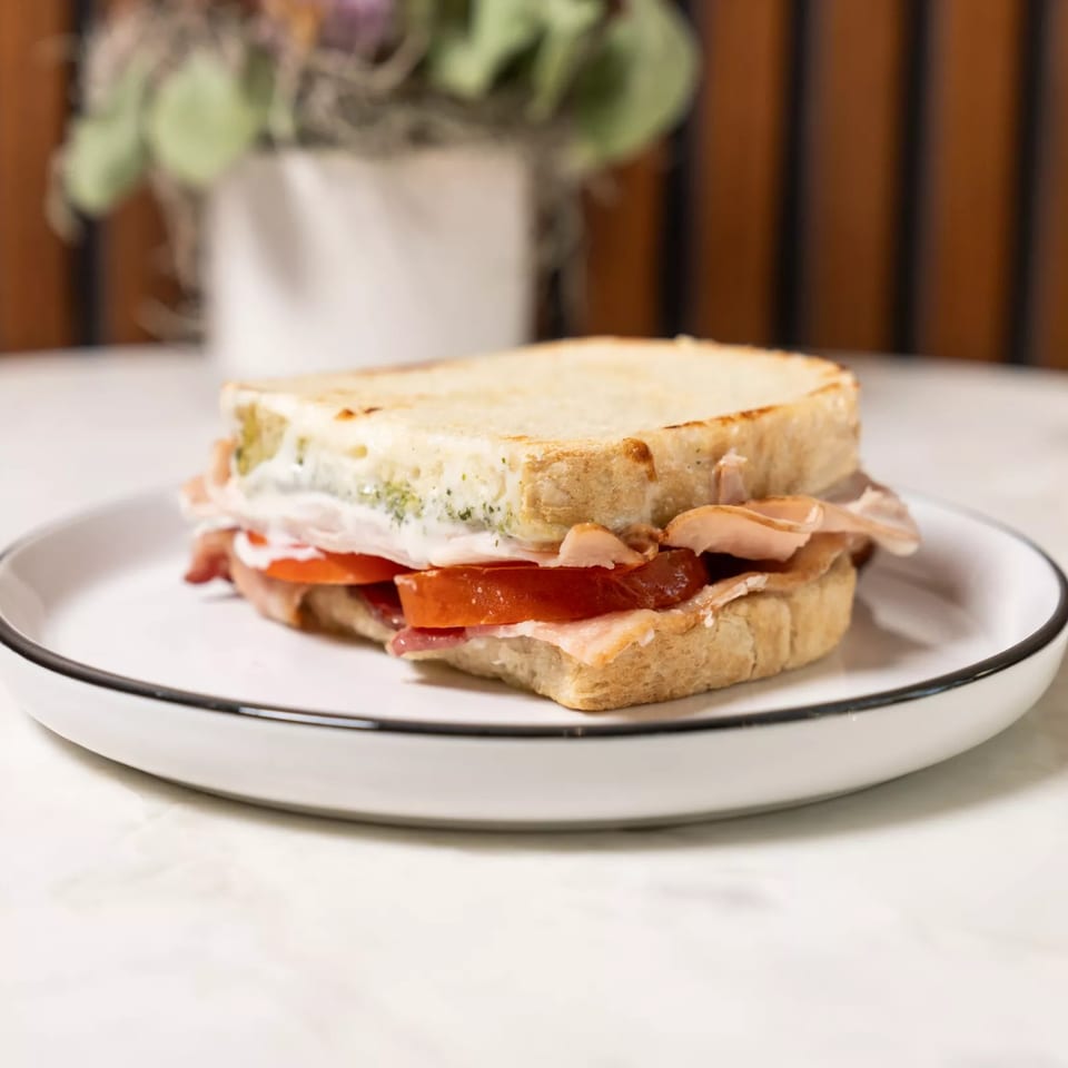 A grilled sandwich with bacon, tomato, and a creamy spread on a white plate.