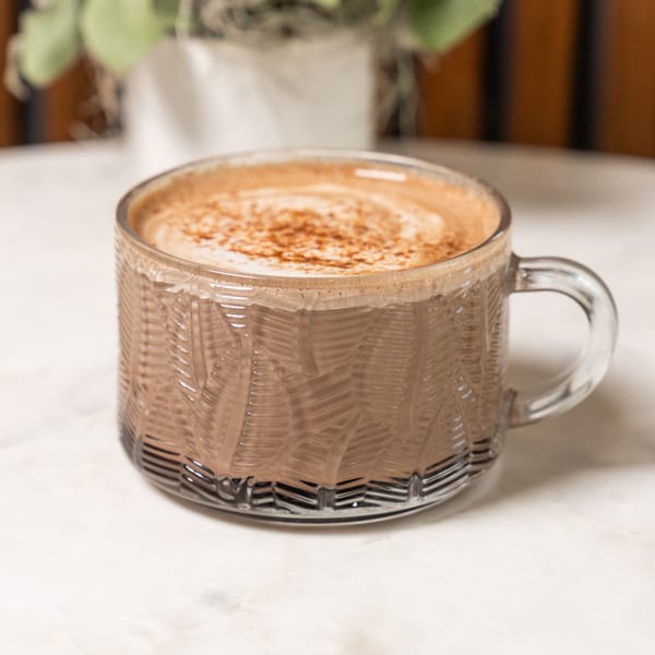 A glass mug filled with a frothy, cinnamon-dusted hot chocolate sits on a white marble table.