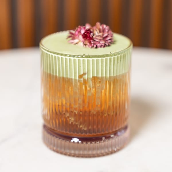 A cocktail with a green foamy top and a pink flower garnish in a textured glass.