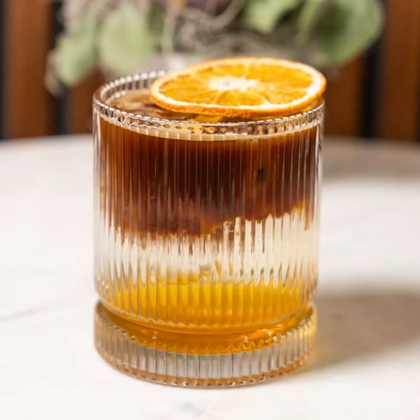 A layered cocktail with dark coffee, white foam, and orange juice, garnished with a dried orange slice.